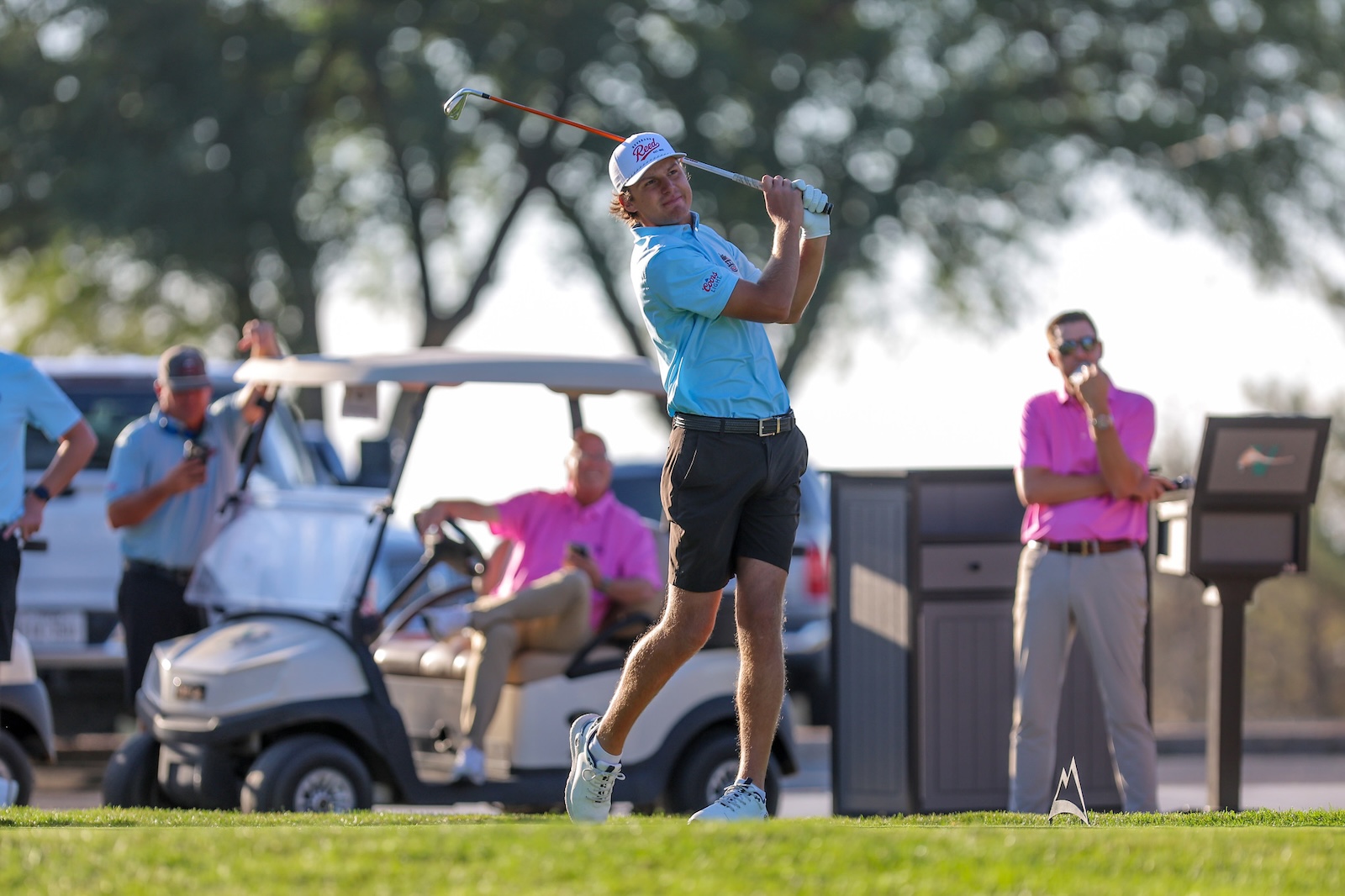 Male golfer in a light blue shirt mid-swing on a sunny golf course, with spectators and a golf cart in the background.