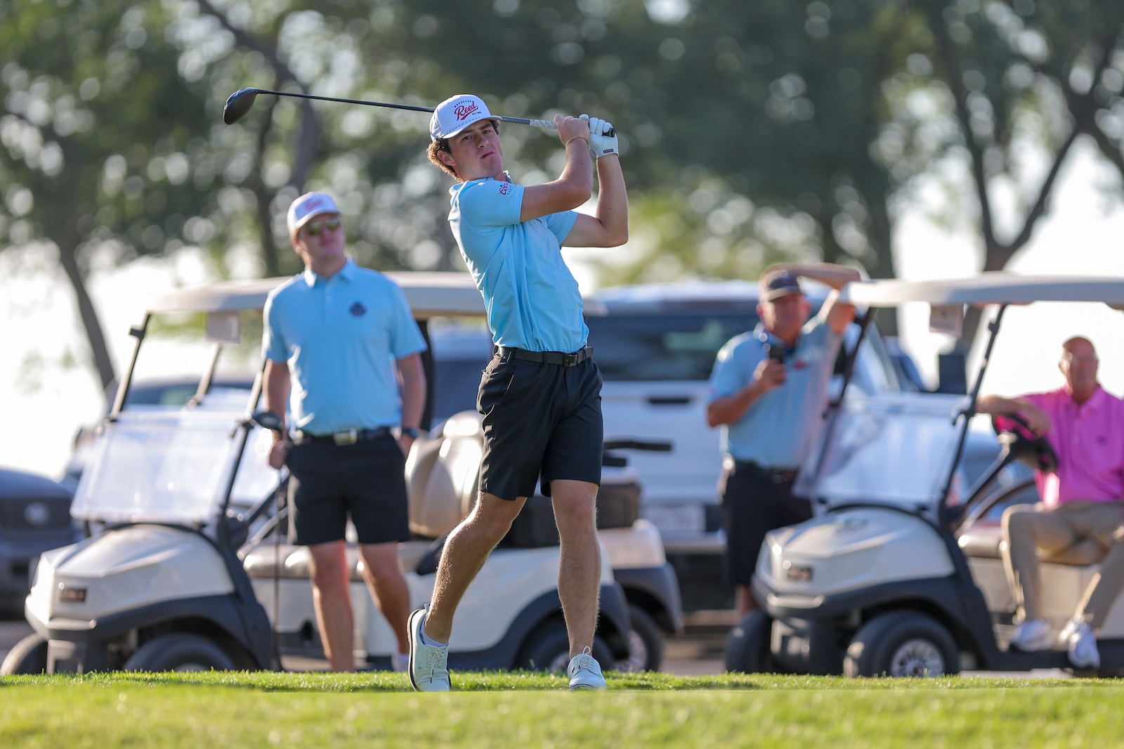 Golfer in light blue shirt mid-swing on a golf course as teammates and golf carts watch in the background.