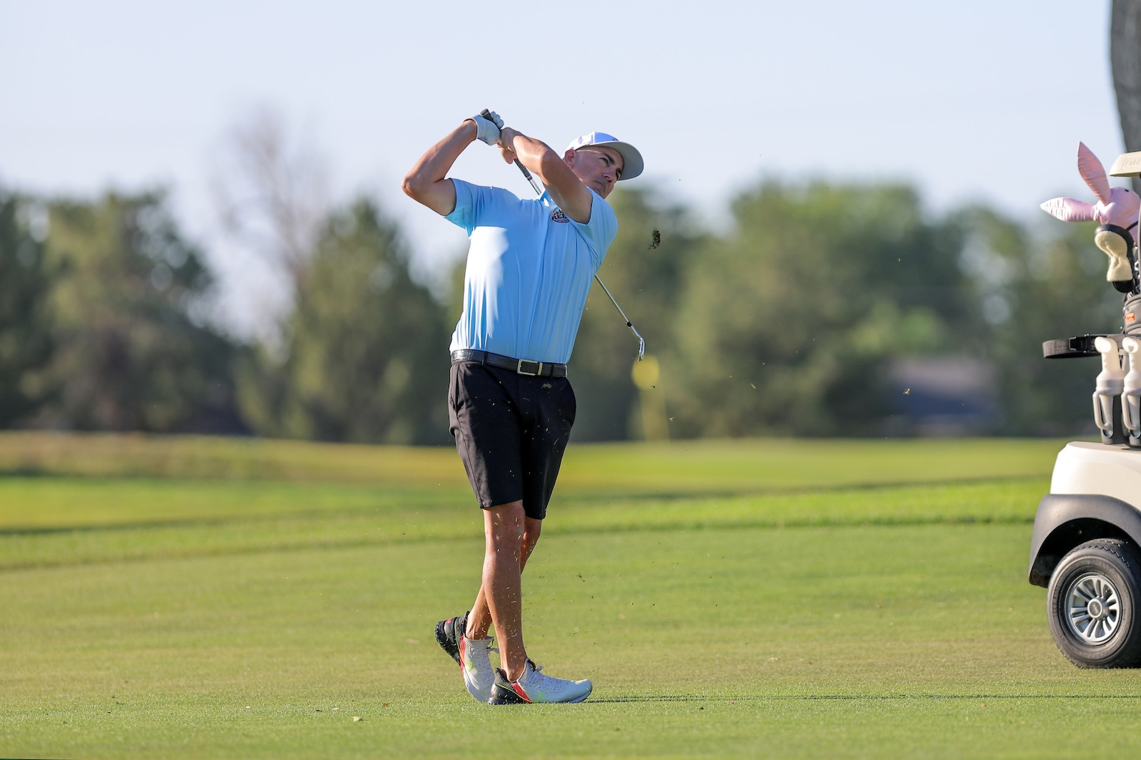 Golfer in a light blue shirt and white cap mid-swing on a sunny golf course; golf cart and bags visible in the background.