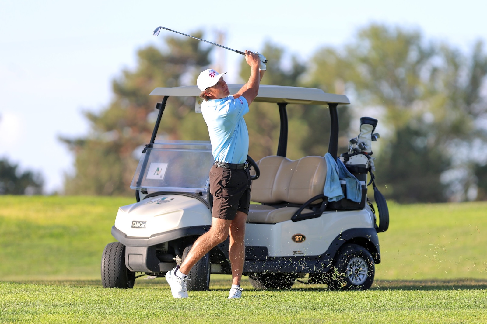 Golfer in a light blue shirt and white cap mid-swing beside a beige golf cart on a sunny course.