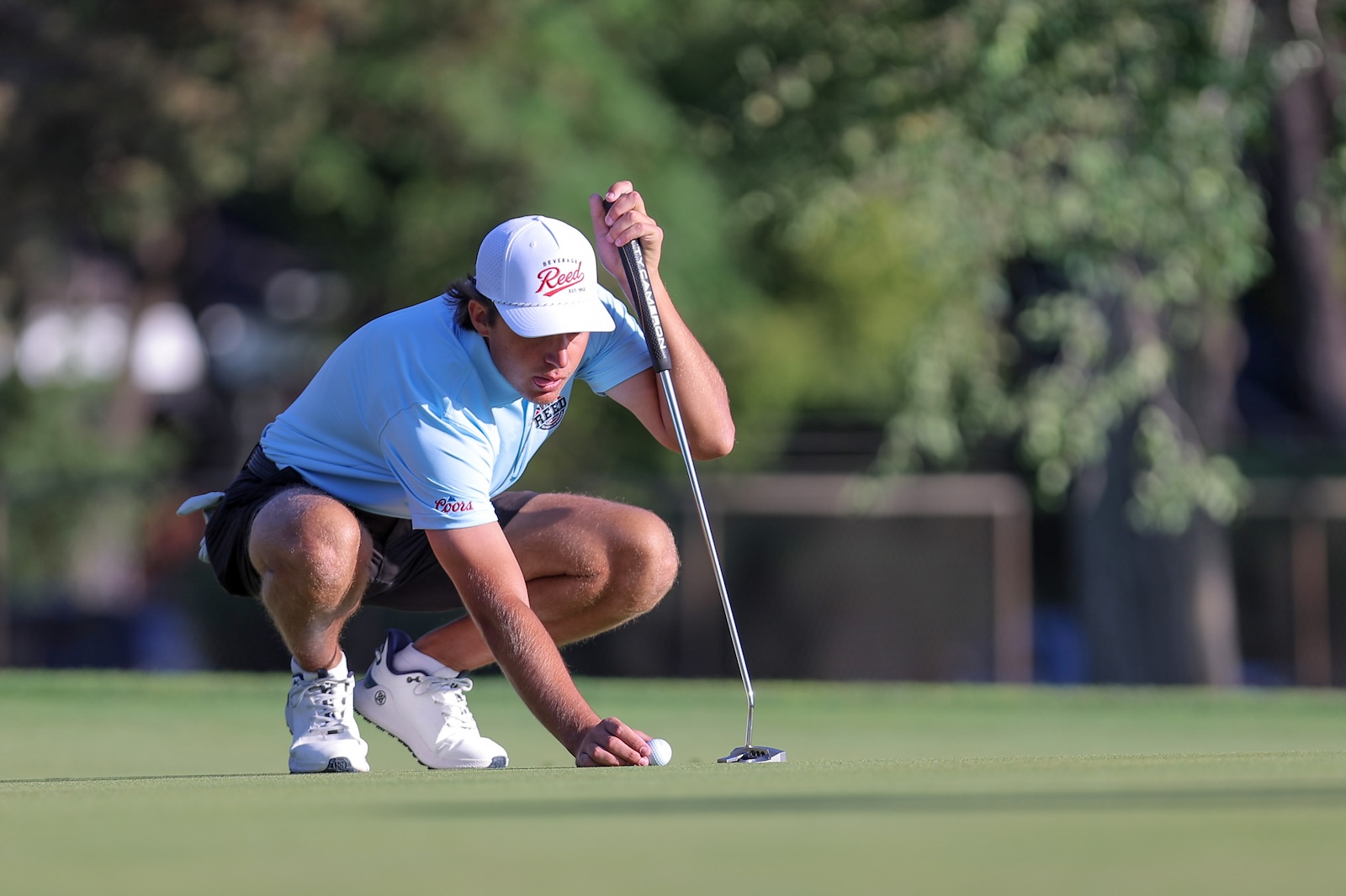 Golfer in a light blue shirt crouches to align a putt on the green, with the ball near the hole.