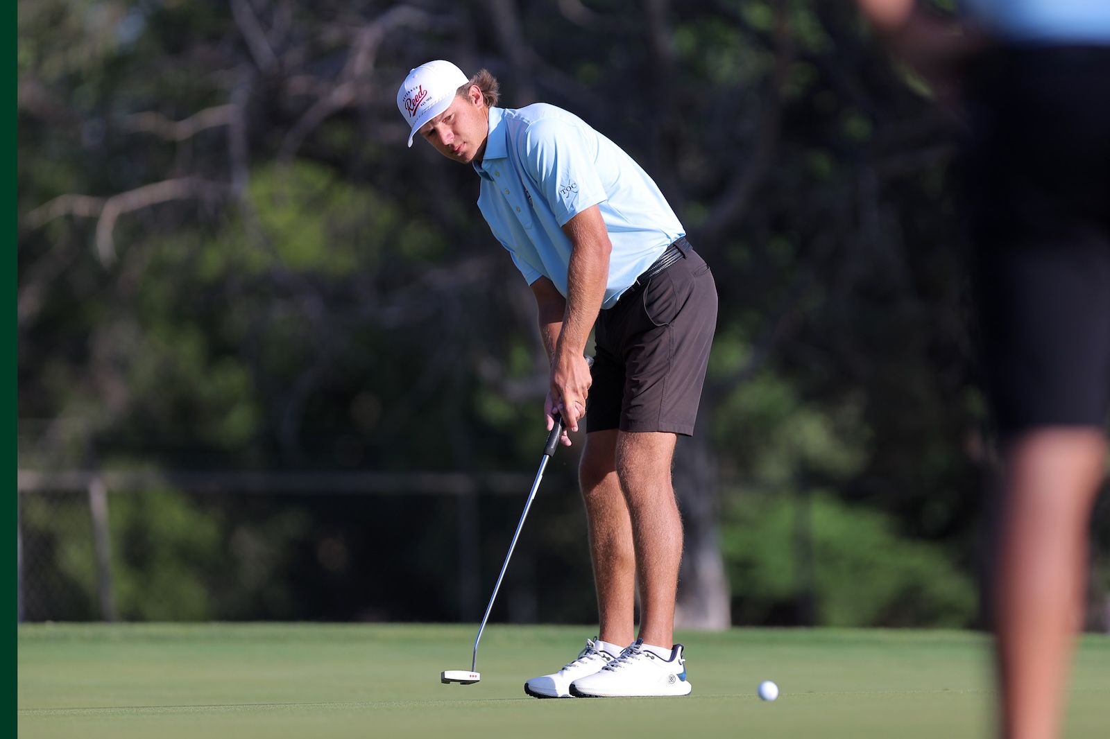 Male golfer lines up a putt on the green, wearing a light blue polo and white cap, focusing on the ball near his club head