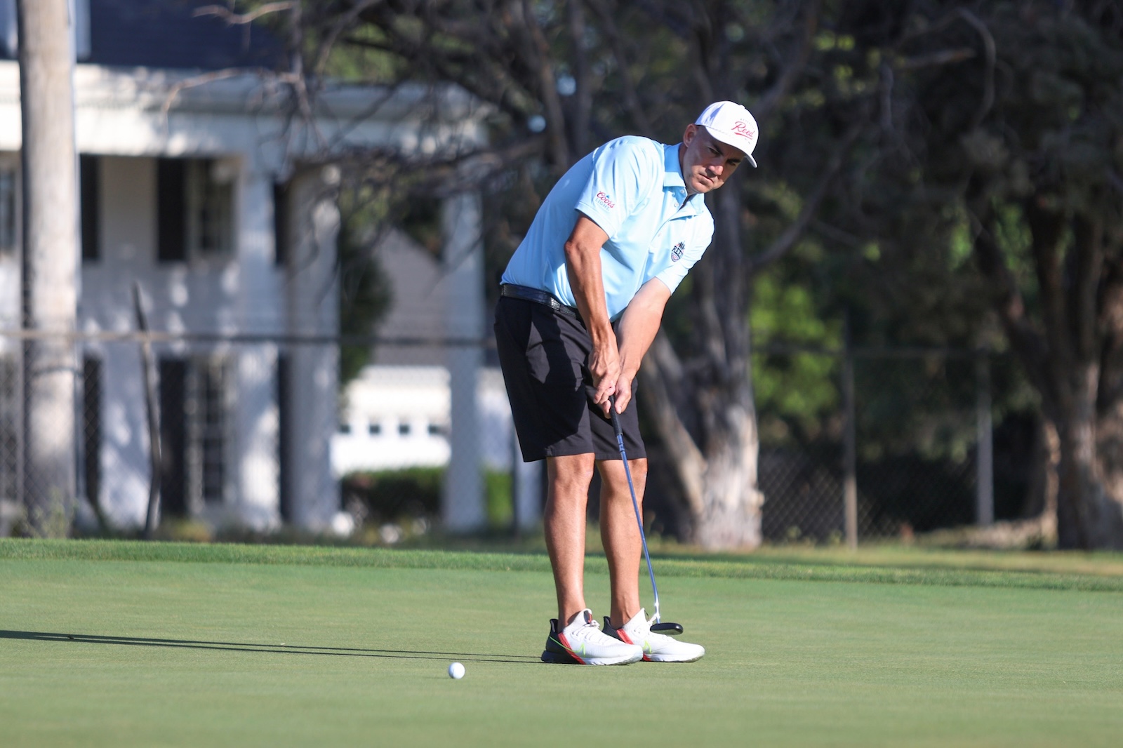 A golfer wearing a light blue polo and white cap lines up a putt on a green, with a nearby ball and putter in view.