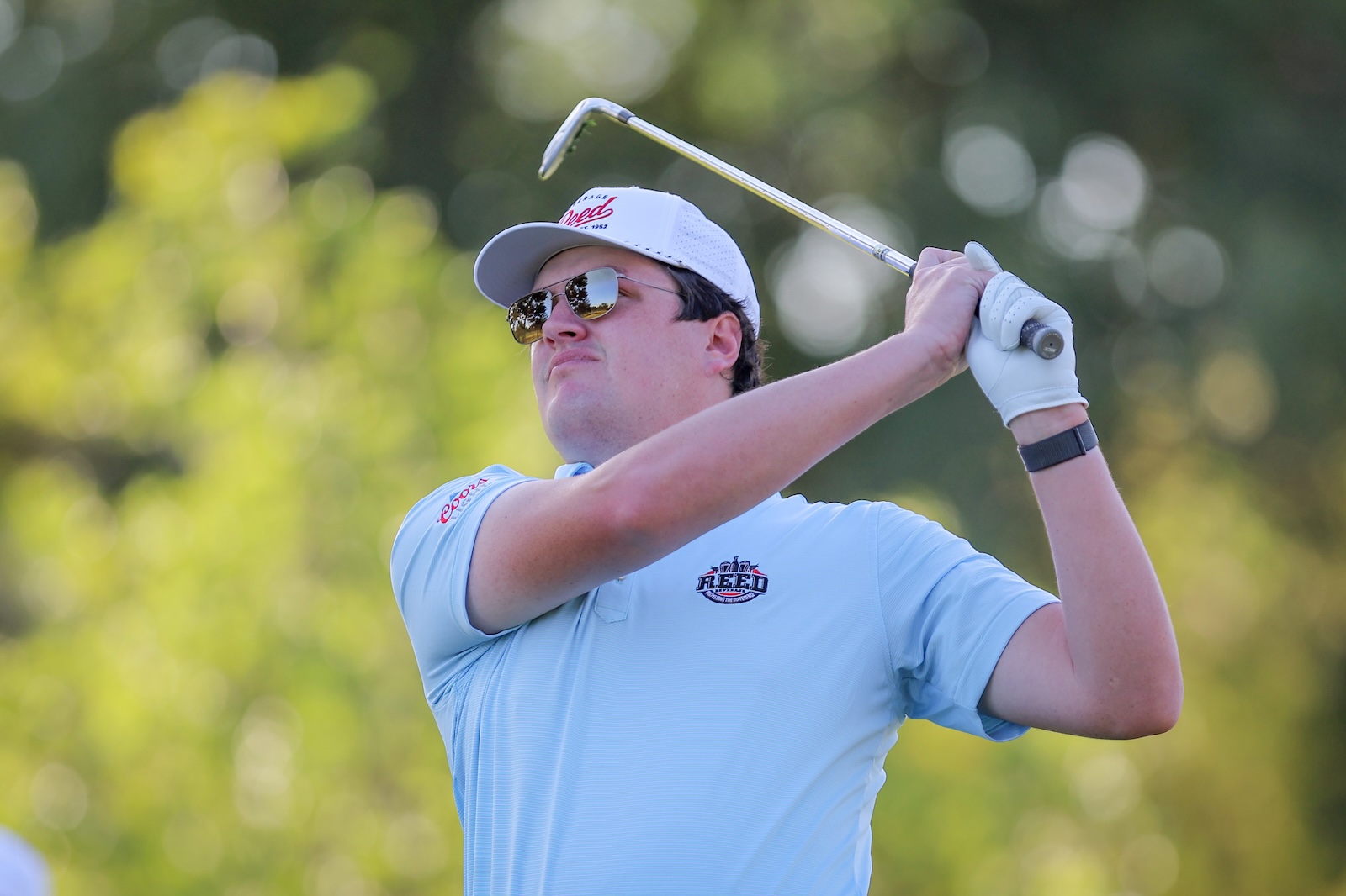 Golfer in a light blue polo wearing a cap and sunglasses, mid-swing on a sunny golf course.