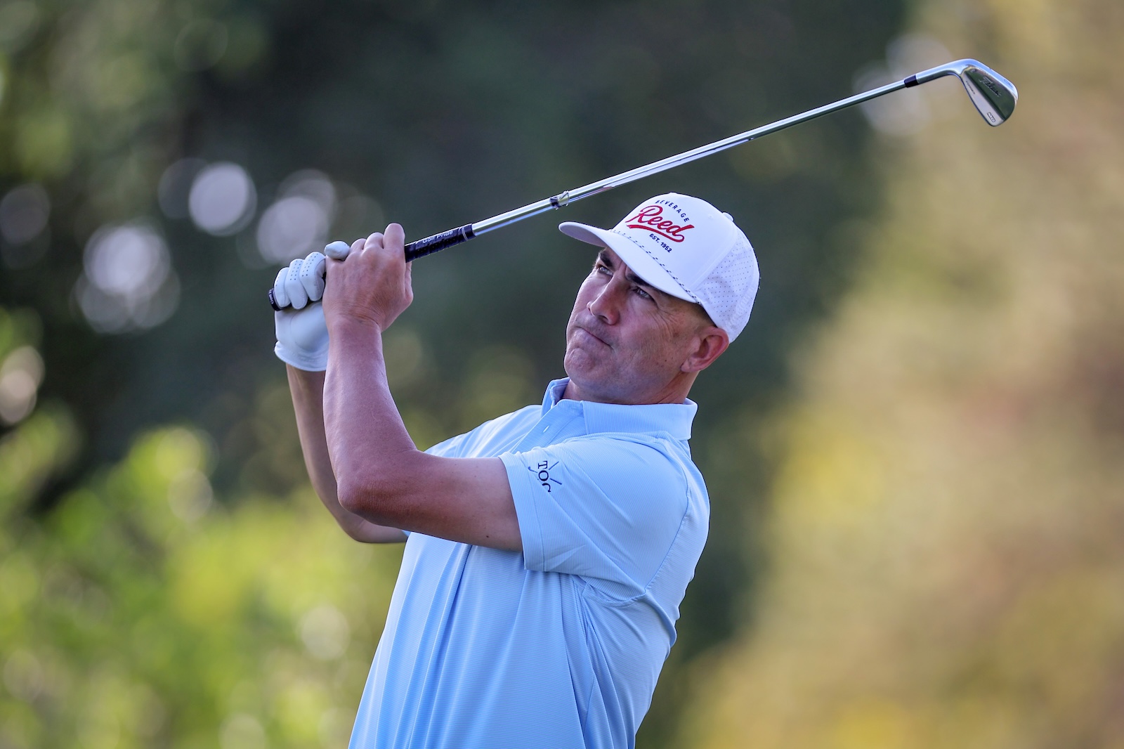 Golfer in a light blue polo and white cap mid-swing on a sunny golf course.