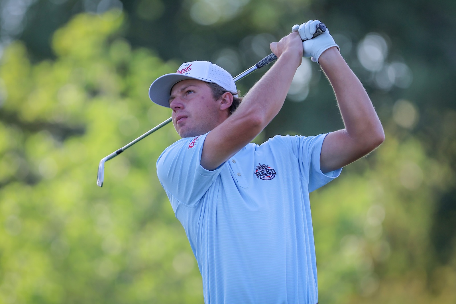 Male golfer in a light blue polo and white cap mid-swing on a sunny course, with a putter in motion and a blurred green background.
