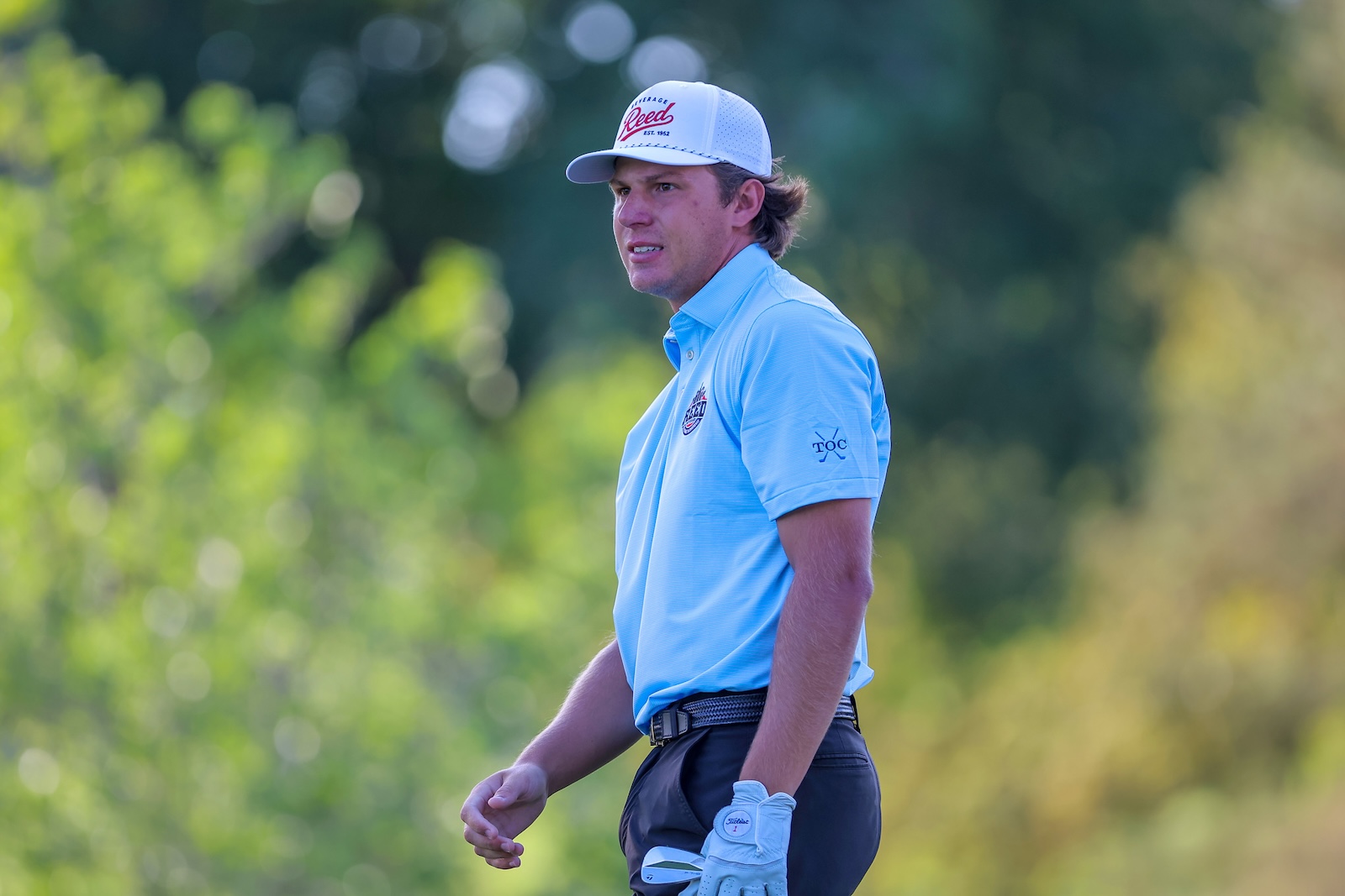 Male golfer standing on a sunny golf course, wearing a light-blue polo and white cap, looking toward the green.