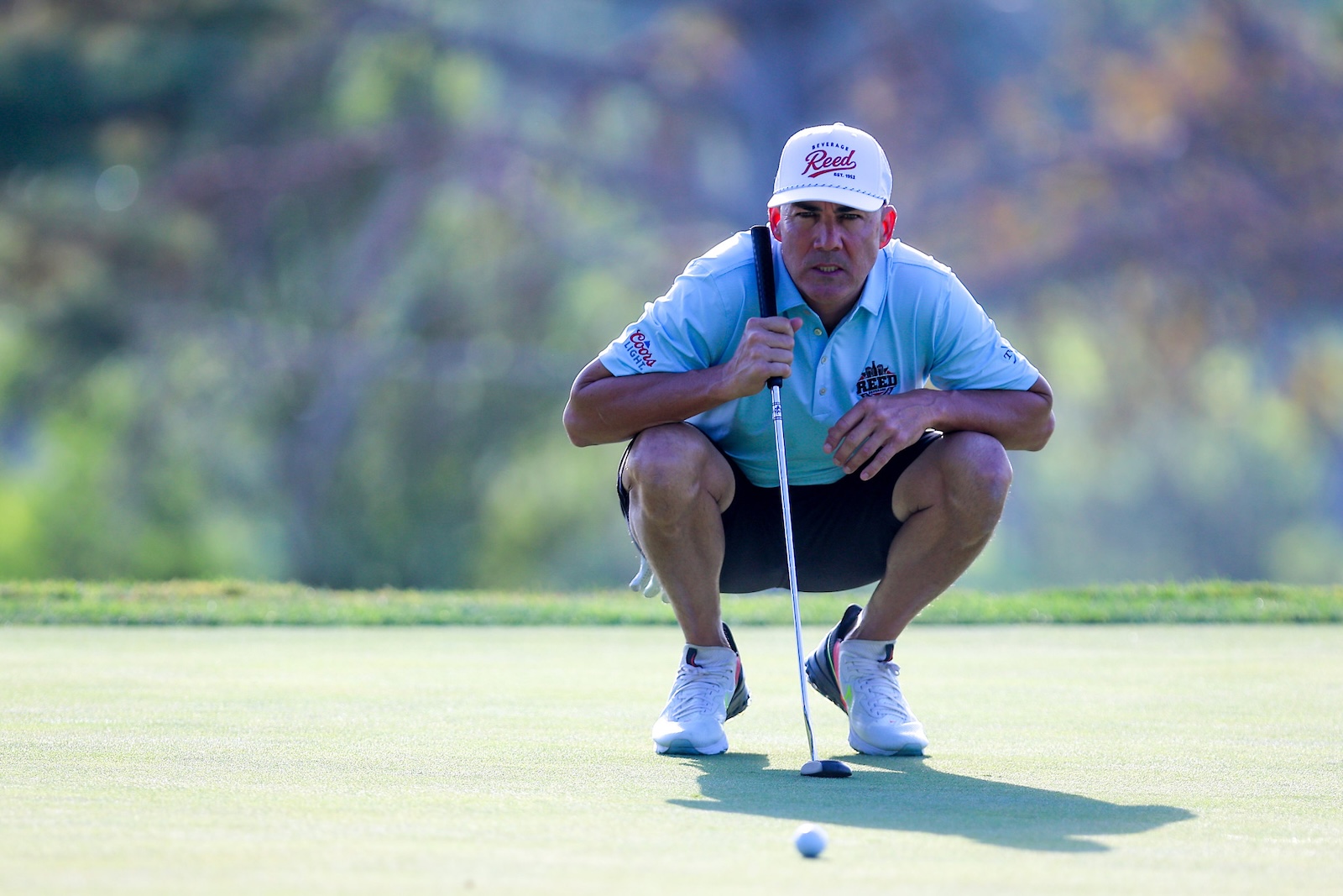 Golfer crouching on a green, lining up a putt with a putter and a ball nearby.