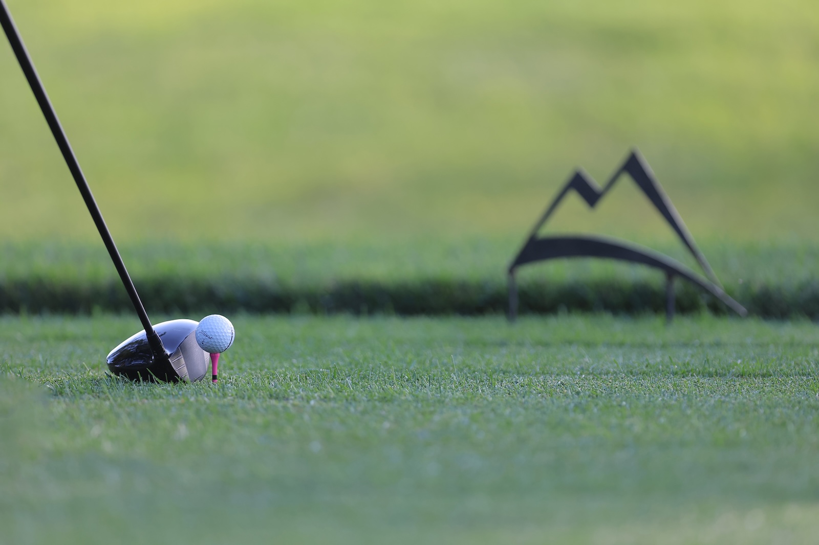 Close-up of a golf driver lining up a white ball on a pink tee on a green fairway, with a blurred backdrop and a distant zigzag flag shape.