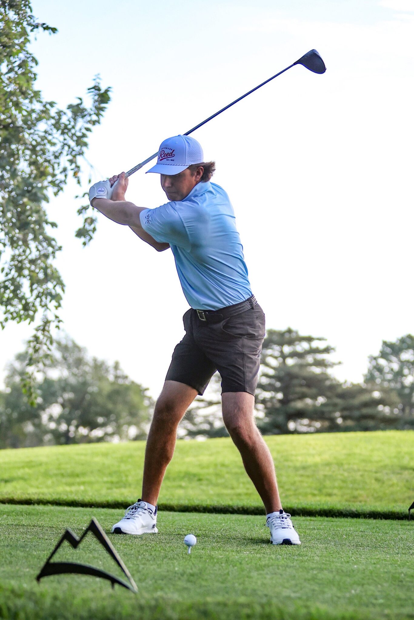 20260813-CoorsTOC-Day1-024 | Press Pass Sports Golfer mid-swing on a sunny course, wearing a blue shirt and white cap, with a tee and ball in the foreground.