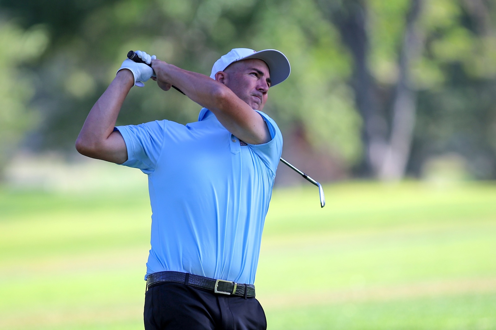 Male golfer in a light blue polo and white cap mid-swing on a sunny golf course.