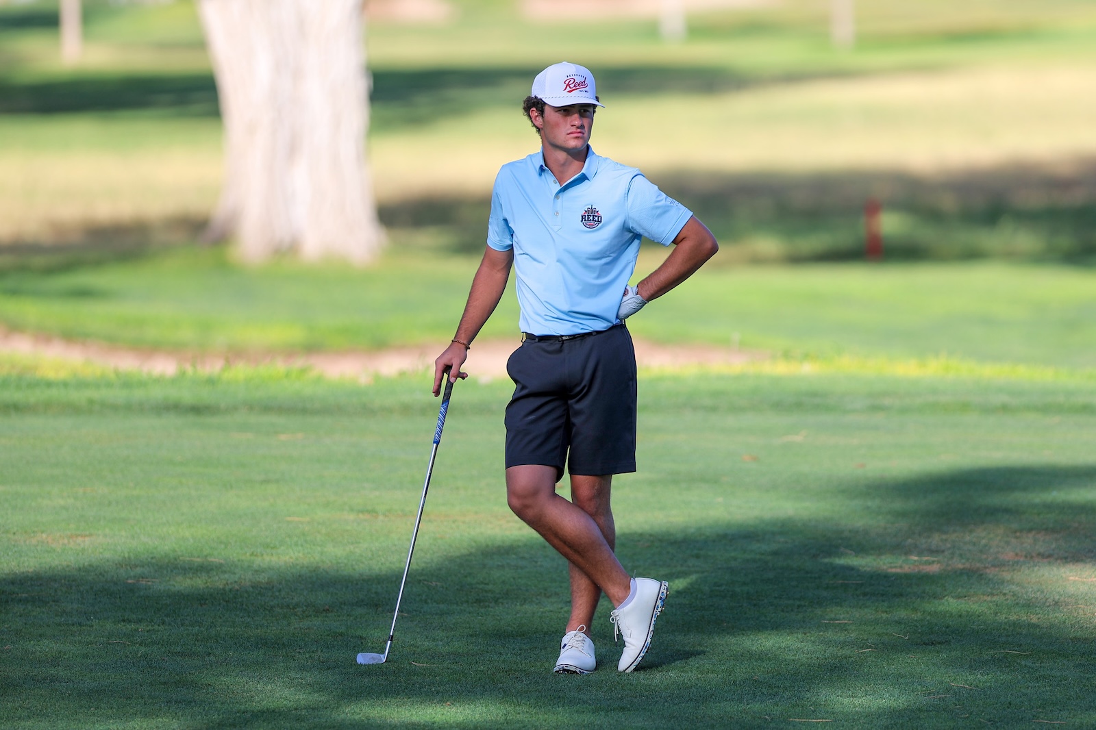 Young male golfer in a light blue polo and white cap stands on a green golf course with a club resting on the ground, looks to the side.