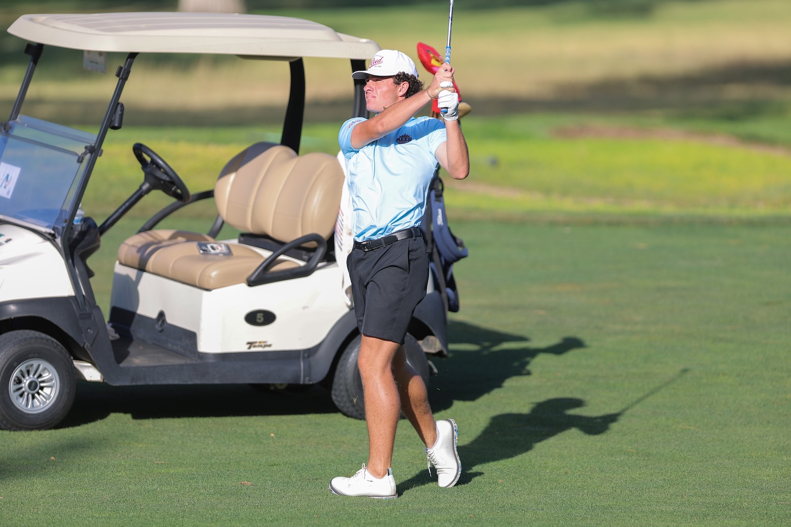 Male golfer in a light blue shirt swings a golf club on a sunny course beside a beige golf cart.