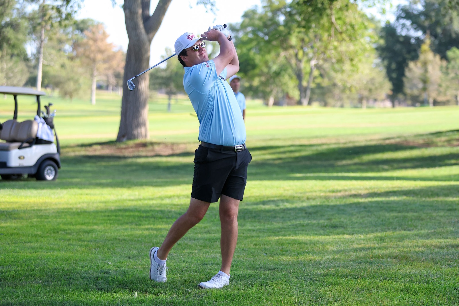 Golfer in a light blue shirt and white cap mid-swing on a sunny golf course, with a golf cart in the background.