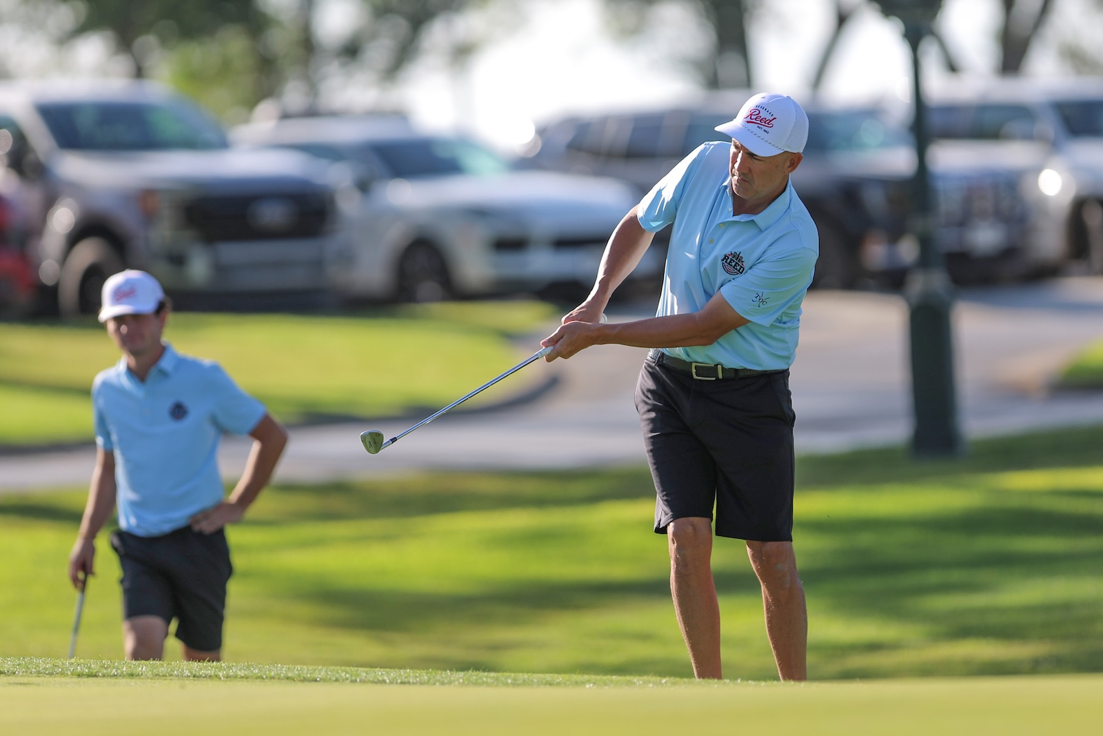 Two golfers in light blue shirts on a sunny golf course; one is mid-swing with a club, another watches in the background, with cars parked beyond the green.