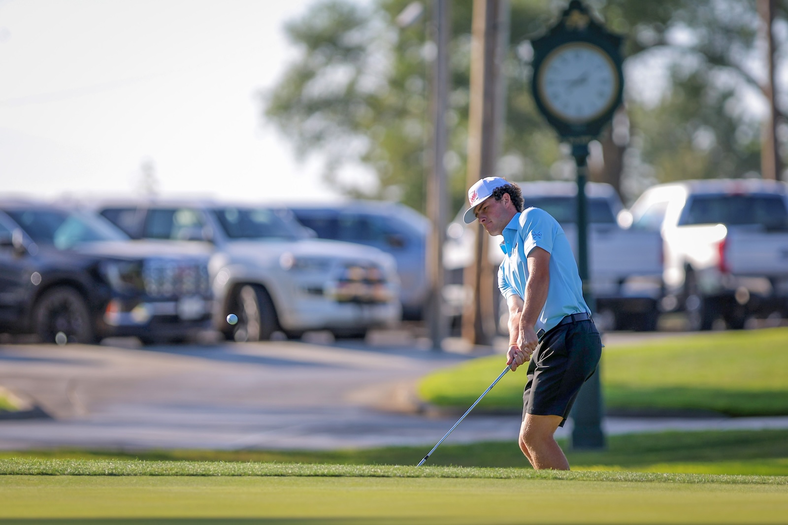 A golfer in a blue polo and cap swings a club on a green, with parked cars and a clock in the background.