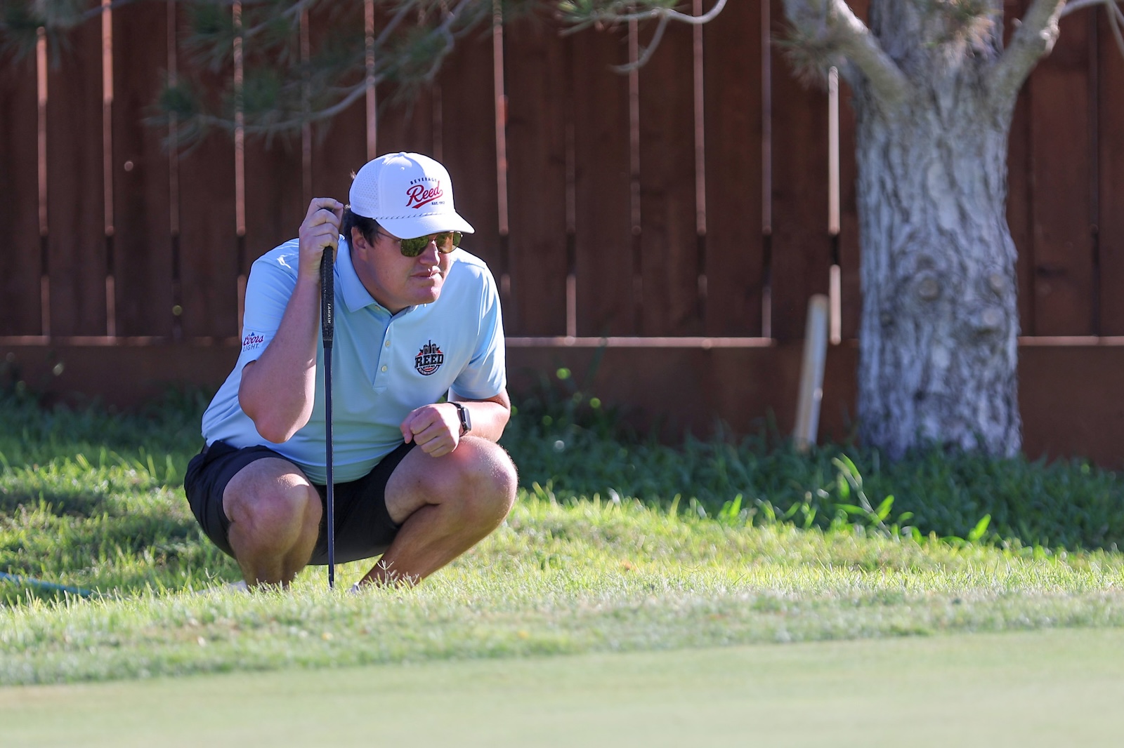 Crouching golfer on a green, wearing a light blue polo, white cap, and sunglasses, holding a putter.