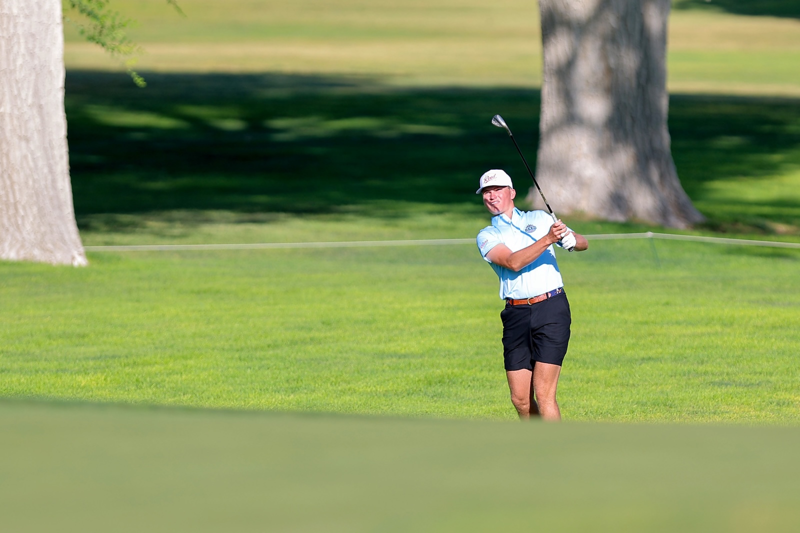 Golfer in a light blue shirt and white cap mid-swing on a green golf course.
