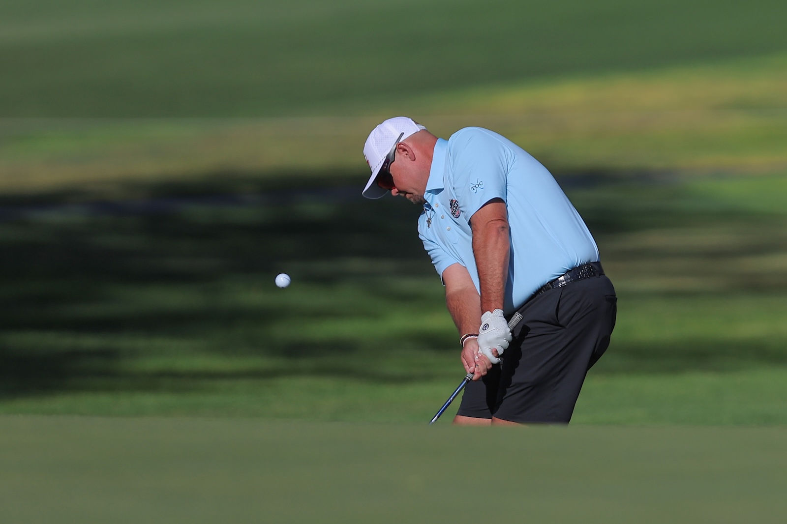 Golfer in a light blue polo and white cap swings on a green golf course as the ball flies through the air.