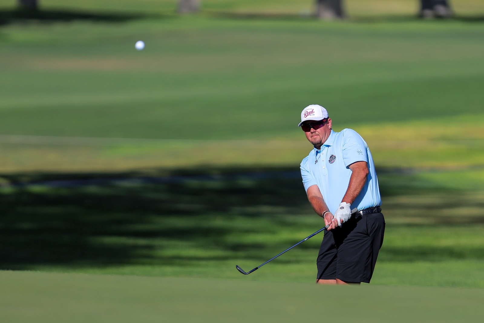 Male golfer in a light blue polo and white cap mid-swing on a green golf course fairway, focusing on his shot