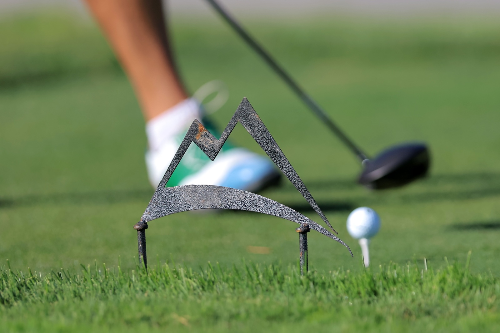 Close-up of a golf ball on a tee with a decorative metal mountain silhouette in the foreground; a golfer's leg and club blurred in the background on the green fairway.
