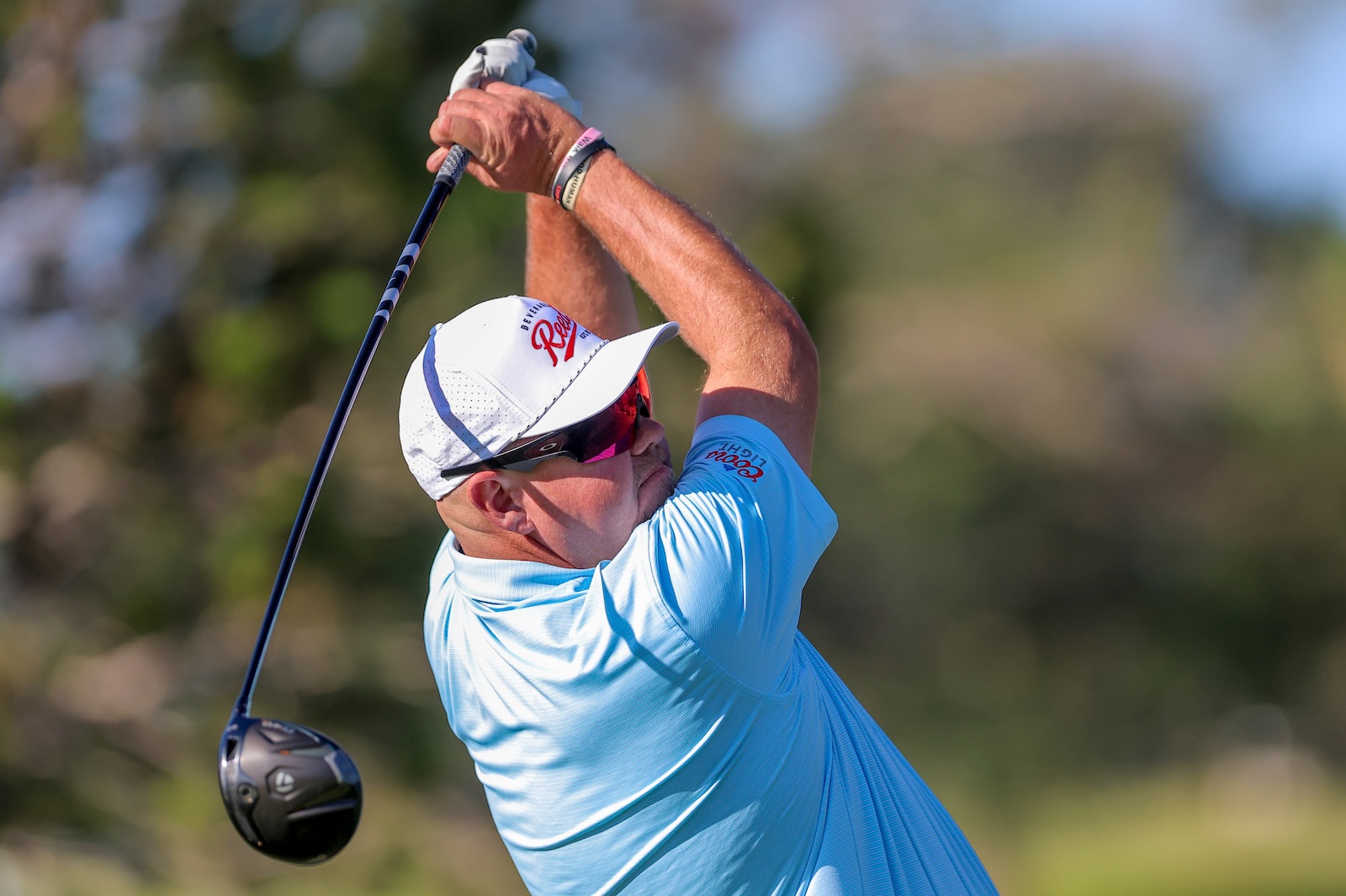 Golfer in a light blue polo swinging a driver, wearing a white cap and sunglasses on a sunny day.