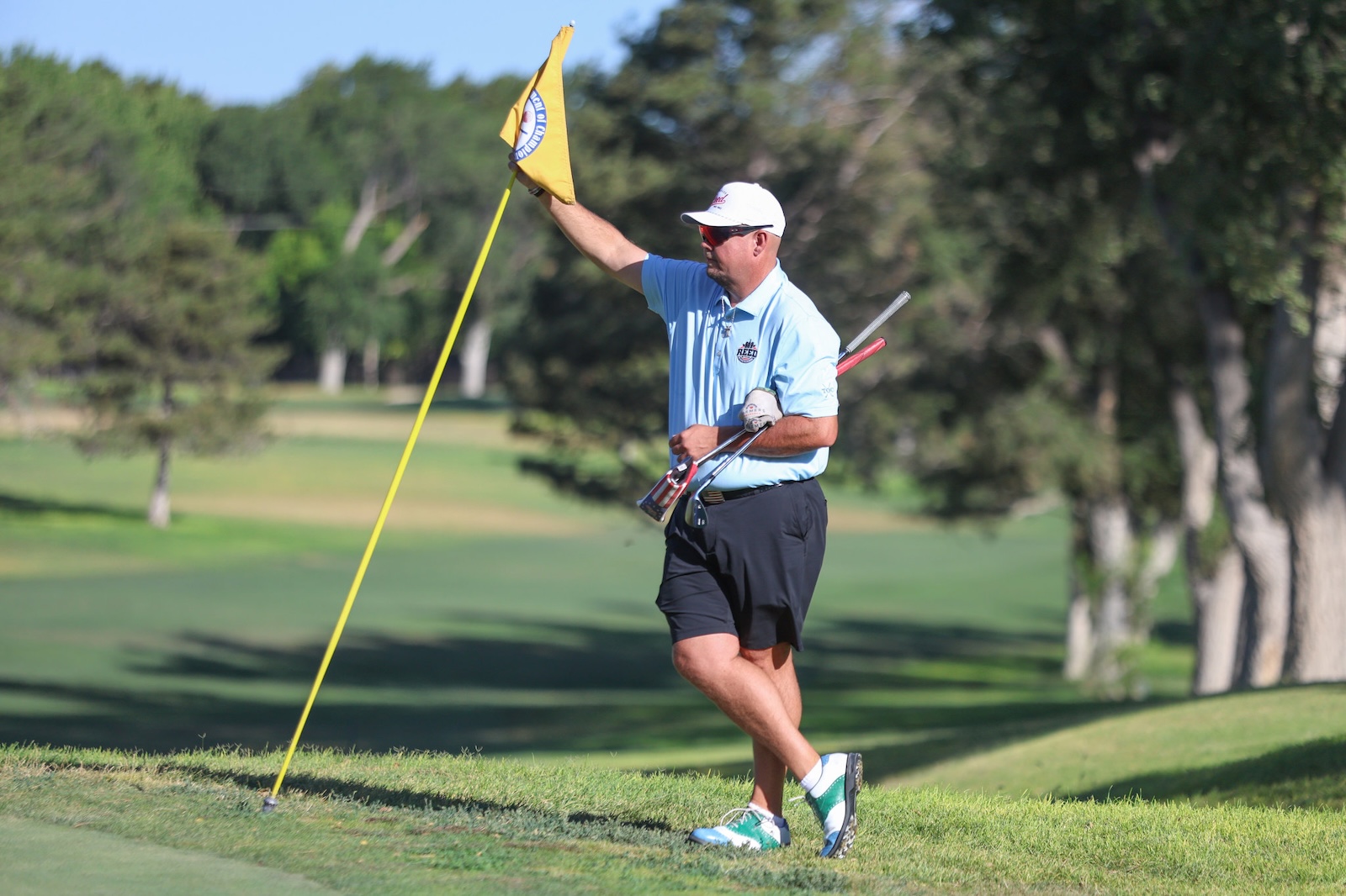 Male golfer in a light blue polo and white cap holds a yellow flag near a hole on a sunny golf course.