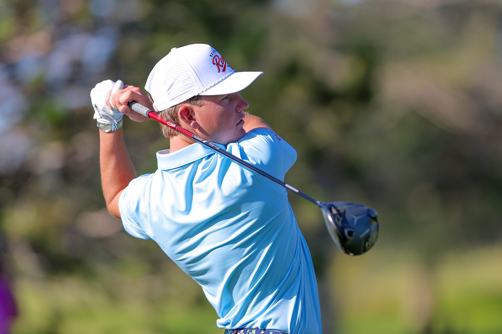 Golfer in a light blue polo and white cap mid-swing with a driver on a sunny course, background blurred.