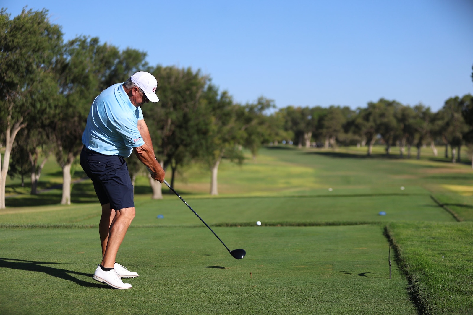 Male golfer in light blue shirt and white cap swings a driver on a sunny golf course, preparing to tee off on a neatly trimmed fairway.