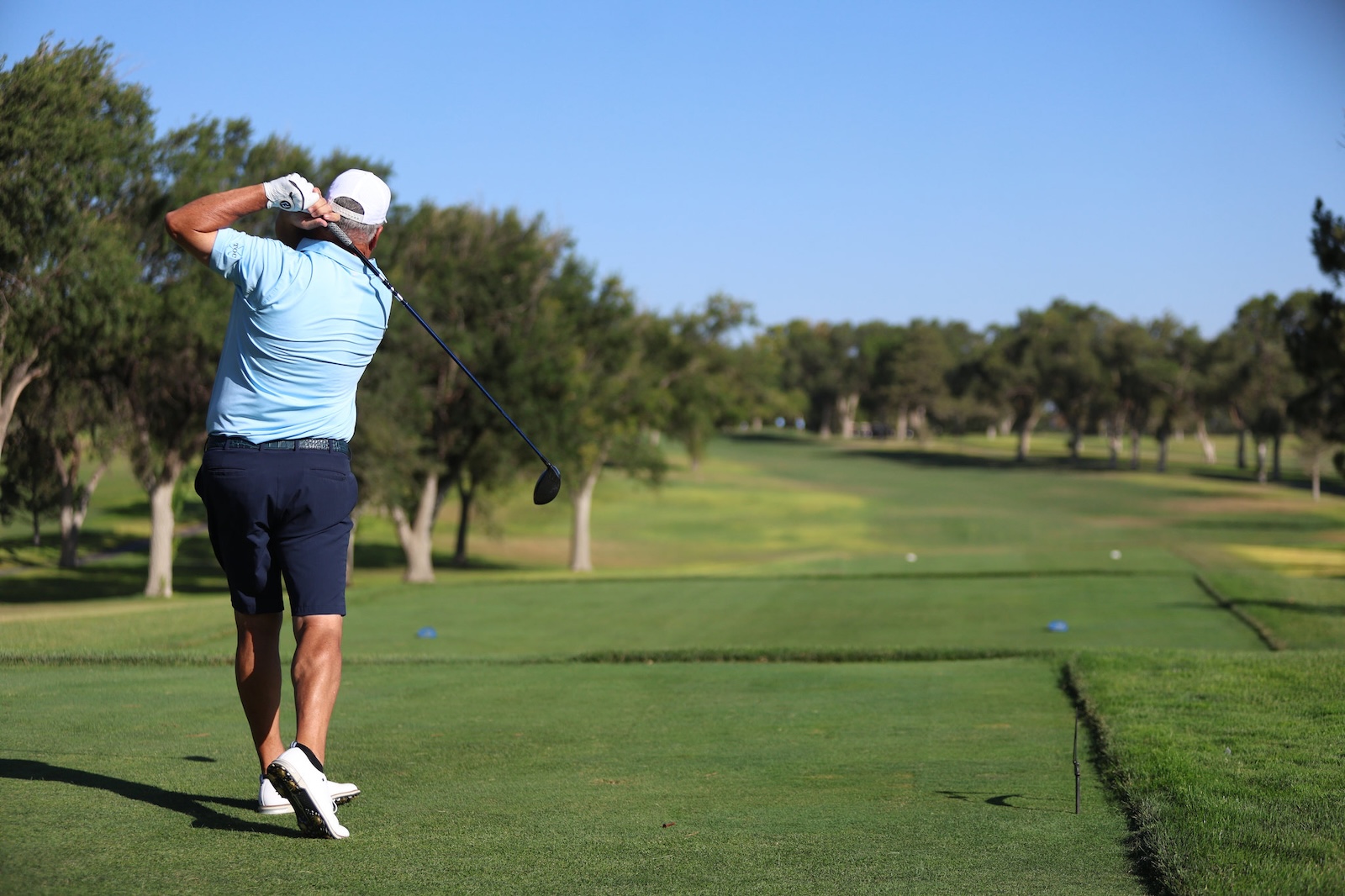 Golfer in a light blue polo and white cap mid-swing on a lush golf fairway under a clear blue sky