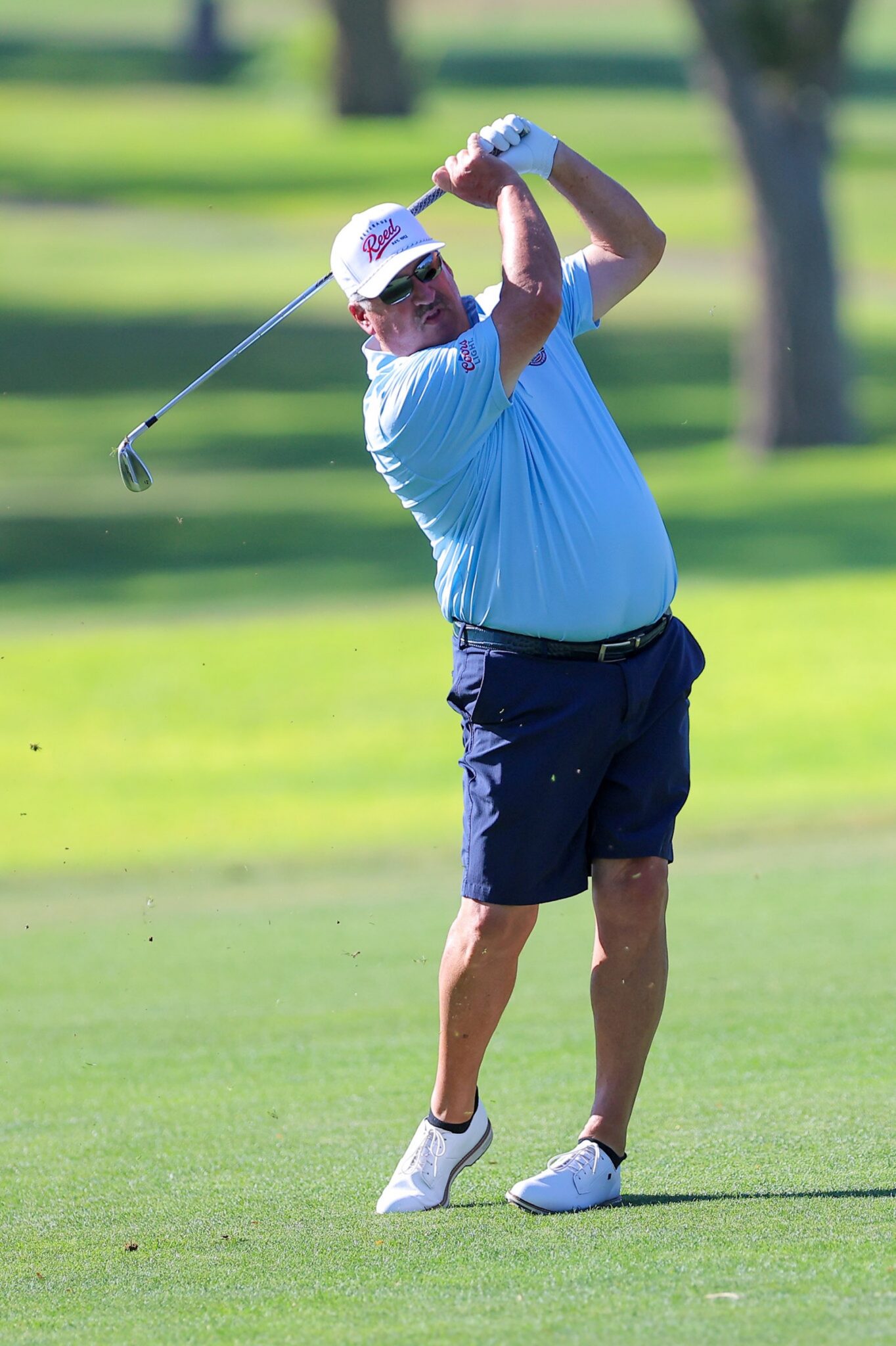 20260813-CoorsTOC-Day1-055 | Press Pass Sports Older man in a light blue polo and navy shorts, swinging a golf club on a sunny green golf course.