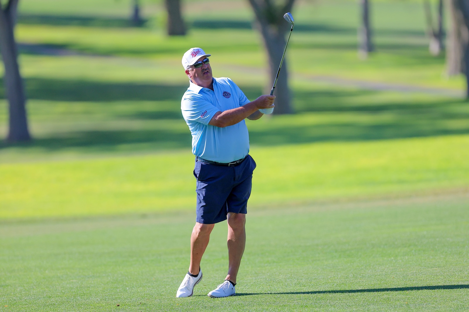 Older man in a light blue polo shirt and navy shorts mid-swing on a sunny golf course, wearing a white cap and sunglasses.