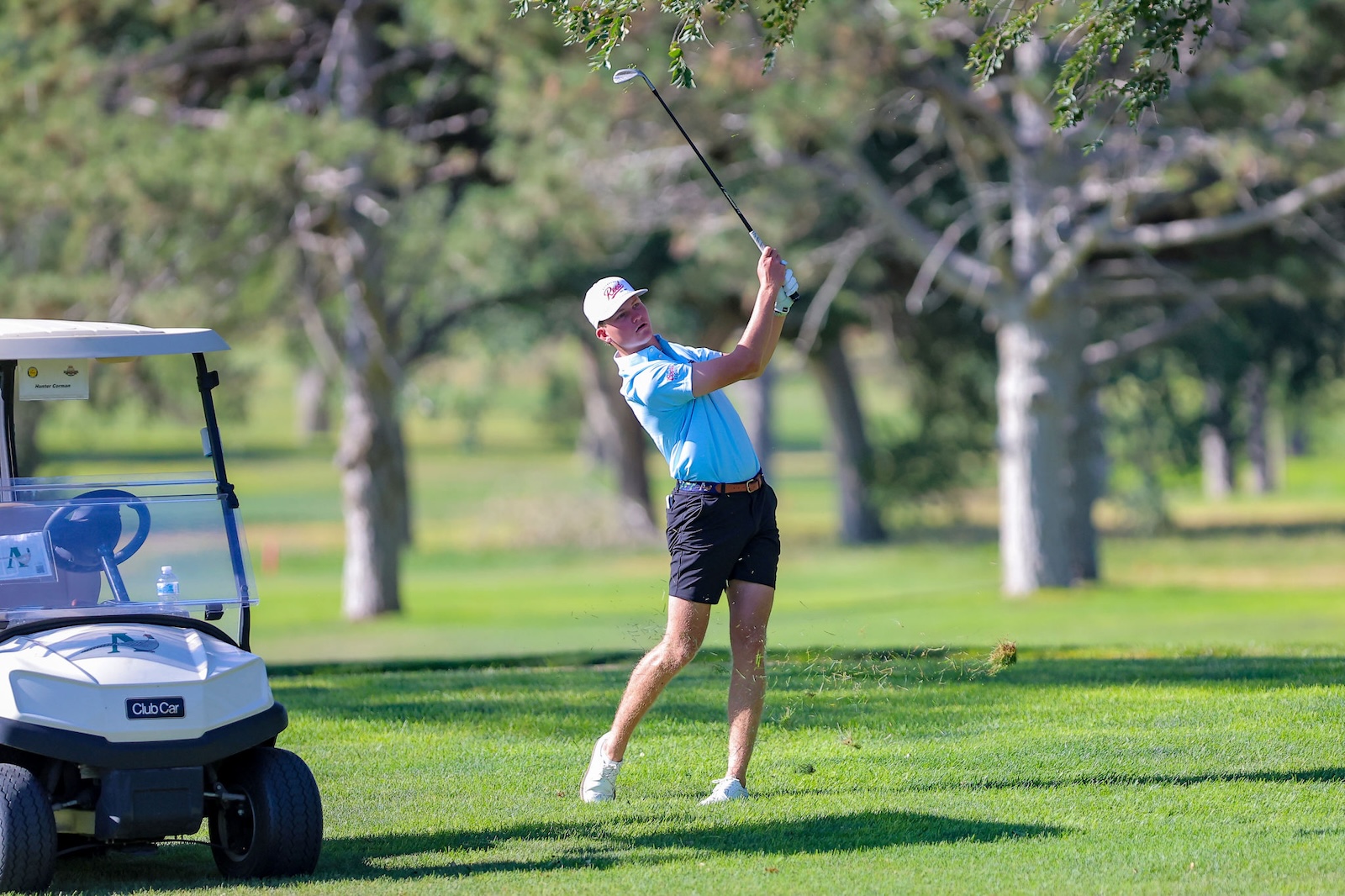 Golfer in a light blue polo and white cap mid-swing on a sunny golf course, with a golf cart nearby on the left.