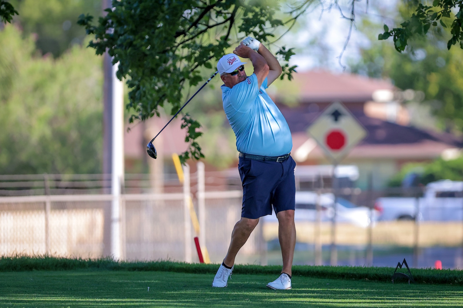 Adult male golfer in a light blue shirt and white cap mid-swing on a sunny course.