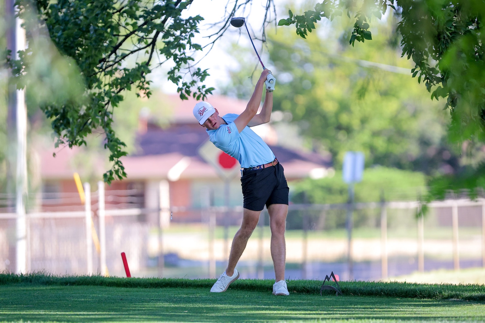 Man golfer in a light blue polo and black shorts teeing off, mid-swing under leafy trees on a sunny course