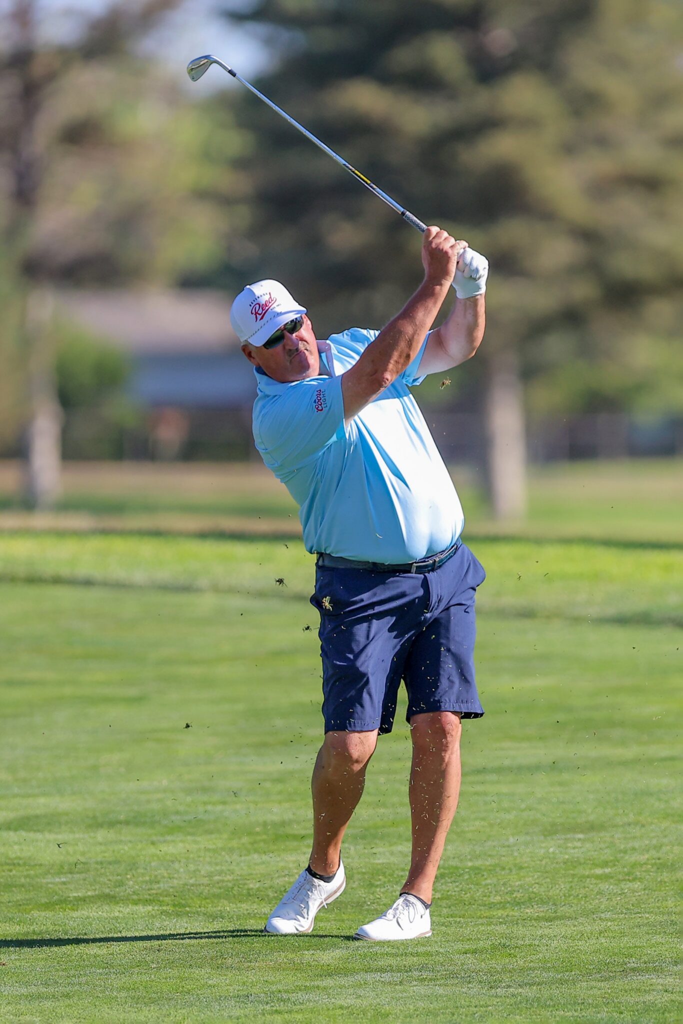 20260813-CoorsTOC-Day1-067 | Press Pass Sports Man in a light blue polo and white cap swinging a golf club on a sunny golf course, mid-backswing with grass flying.