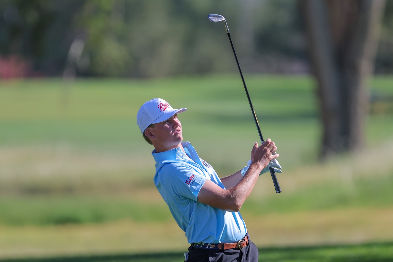 Golfer mid-swing on a sunny golf course, wearing a light blue polo and white cap.
