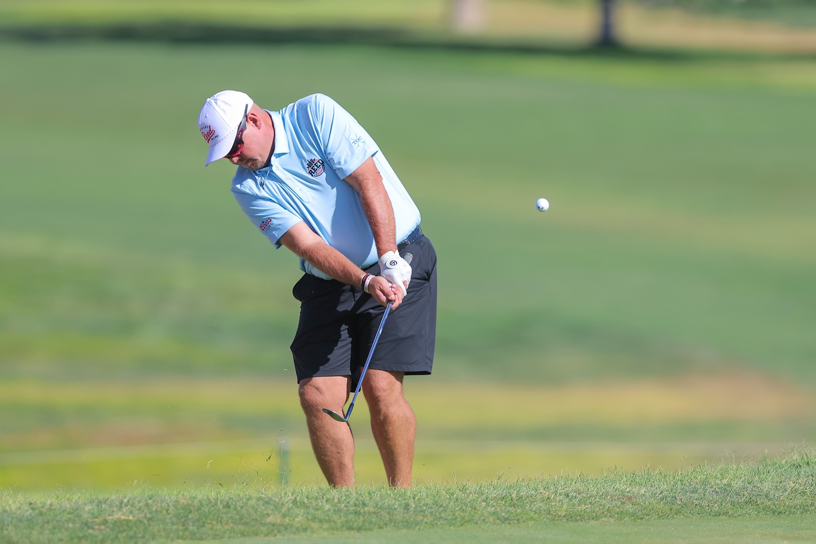Male golfer in a light blue polo and white cap swinging a club on a green fairway; ball is in midair after contact.