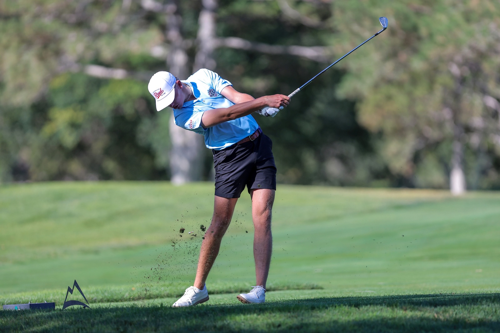 Golfer in a light blue shirt and white cap mid-swing on a lush golf course, dirt flying from his tee box.