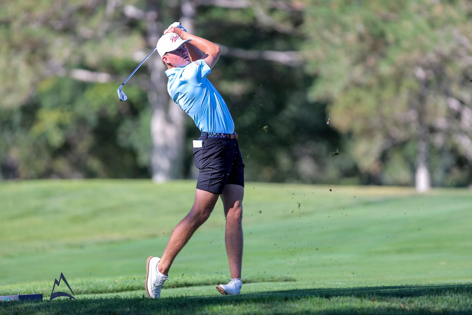 Male golfer in a light blue polo swings a driver on a green golf course; ball just launched into the air behind him.