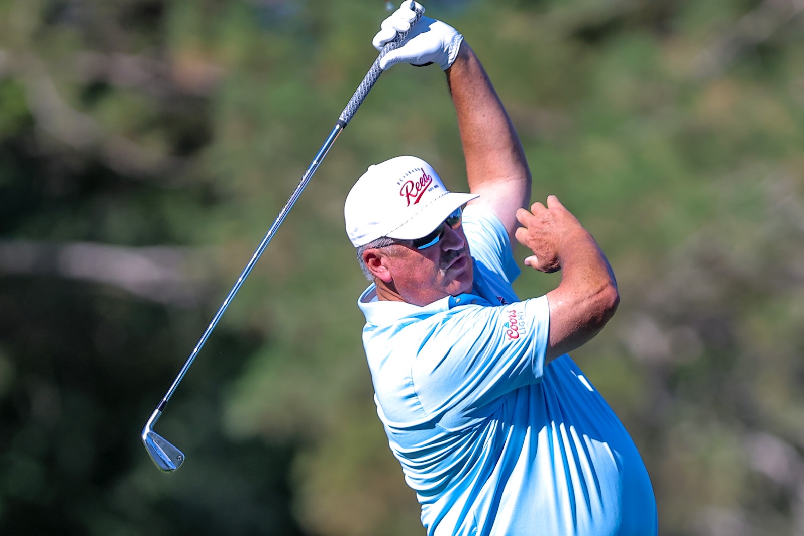 Golfer in a light blue polo and white cap mid-swing on a sunny golf course, with a gloved hand gripping the club.