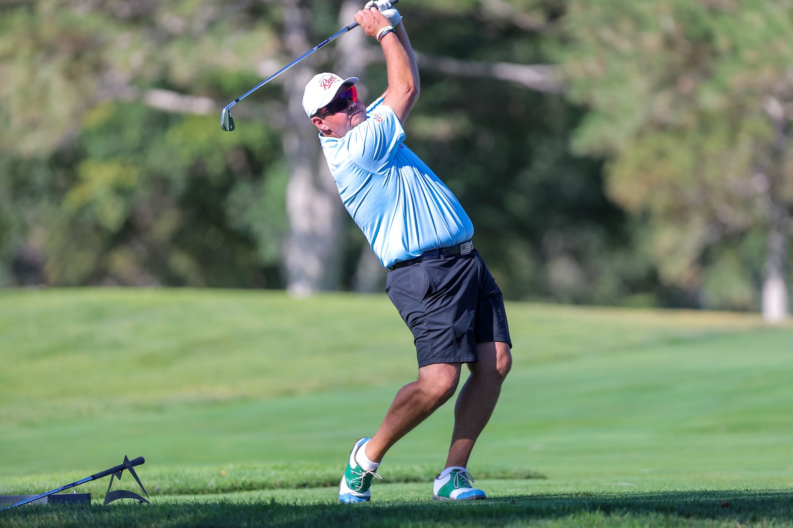 Male golfer in a light blue polo and white cap mid-swing on a sunny golf course.