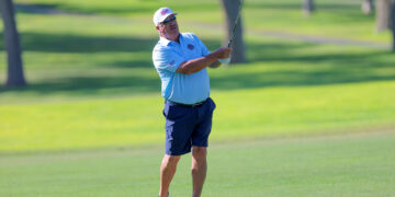 A man in a light blue polo and navy shorts swings a golf club on a lush green course, wearing a white cap and sunglasses.