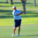 A man in a light blue polo and navy shorts swings a golf club on a lush green course, wearing a white cap and sunglasses.