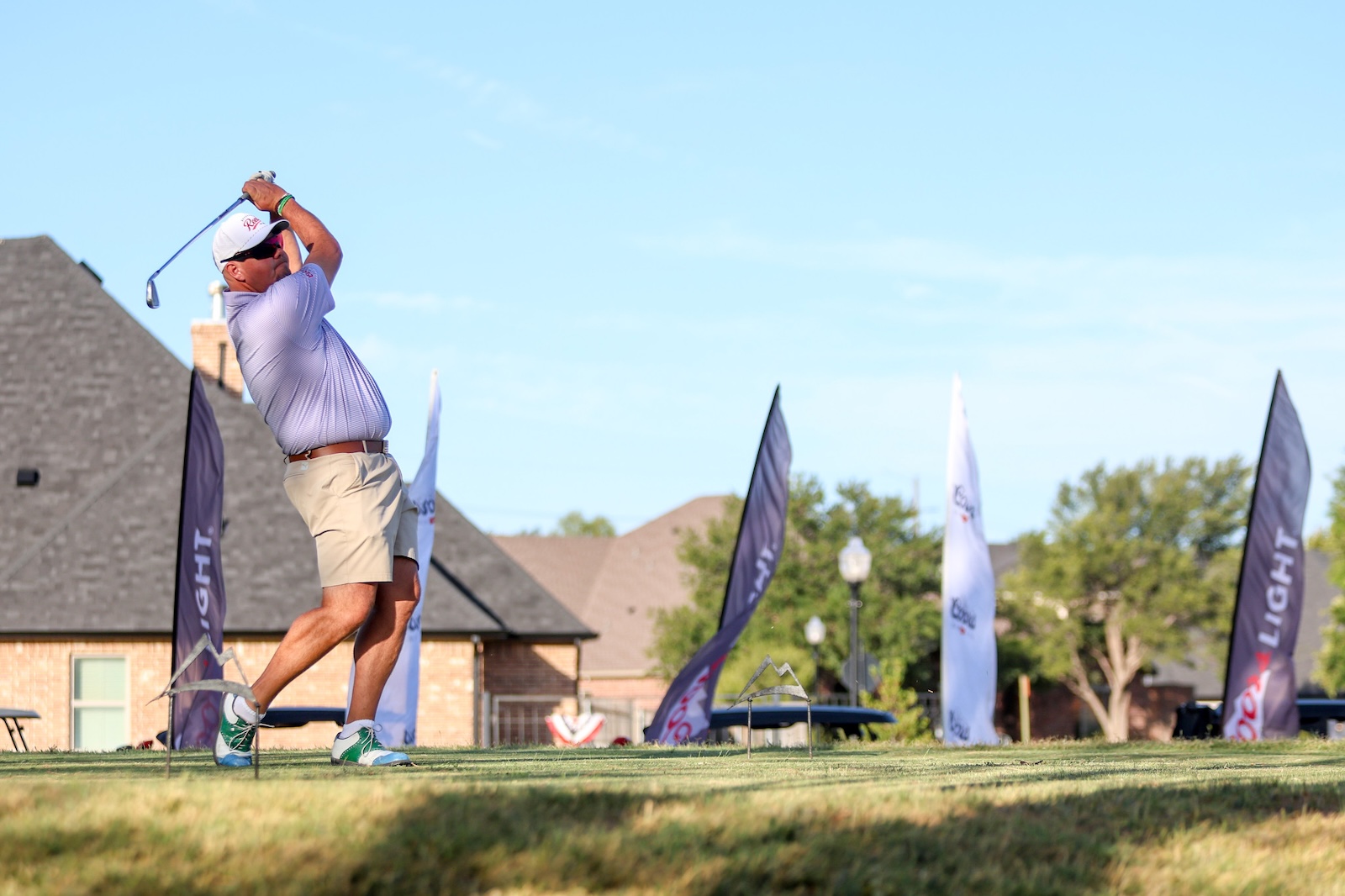Golfer in a light purple polo and beige shorts mid-swing on a sunny golf course with flags in the background.