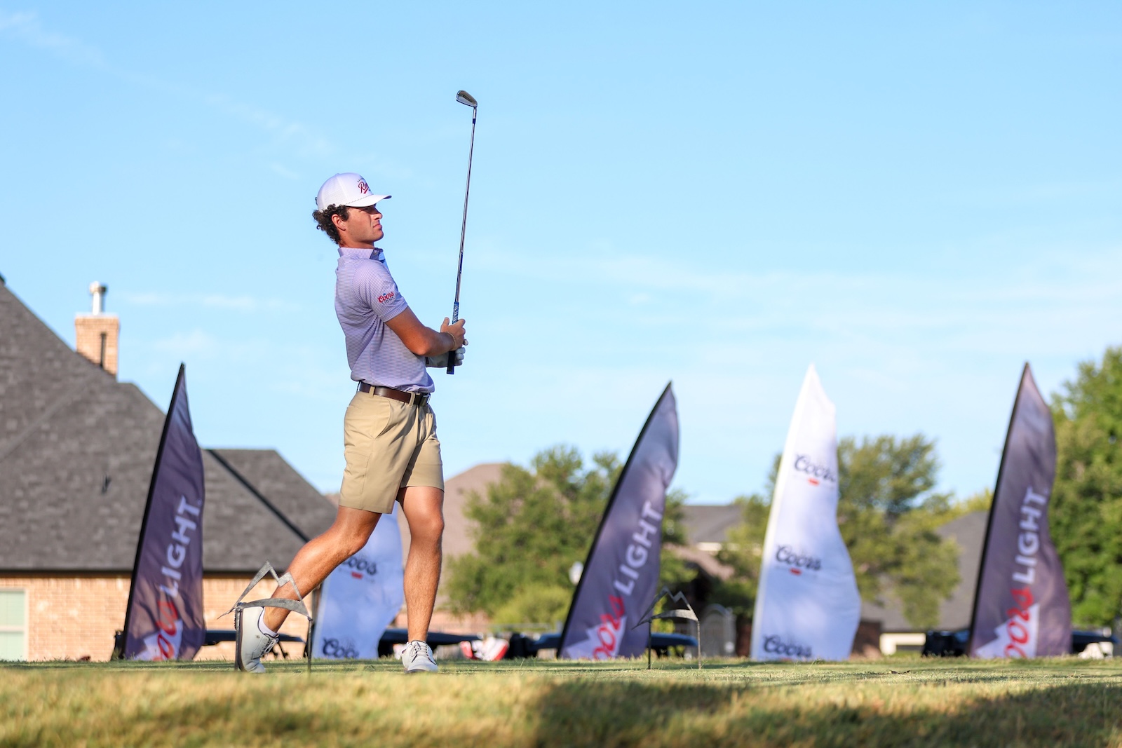 Golfer mid-swing on a practice field, wearing a white cap and beige shorts with banners in the background.