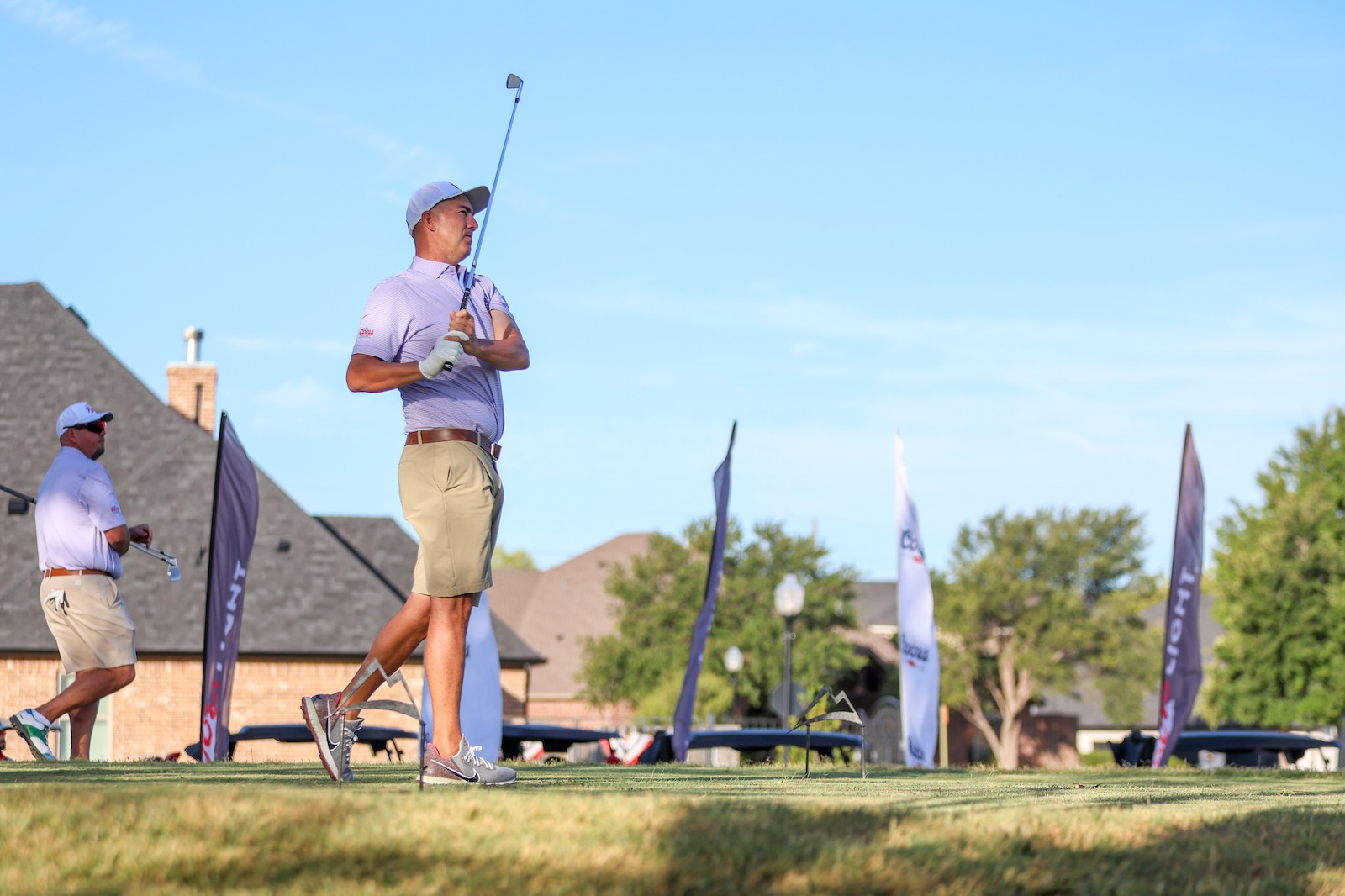 Golfer in lavender polo and beige shorts mid-swing on a sunny golf course with a spectator in the background.