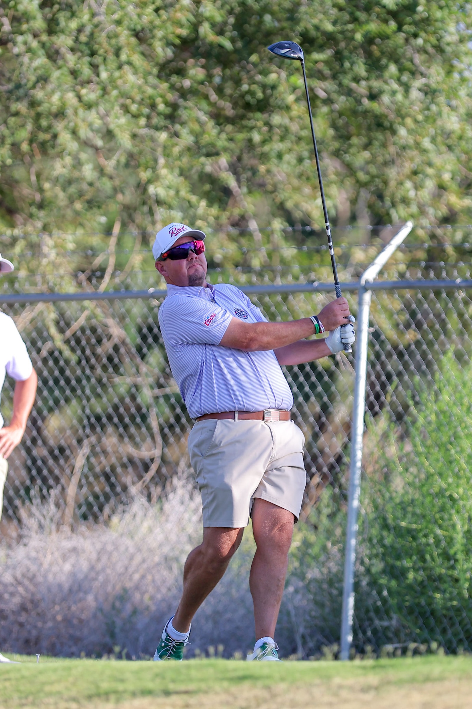 Golfer mid-swing on a golf course, wearing a white cap, red sunglasses, striped polo, beige shorts, and green shoes.