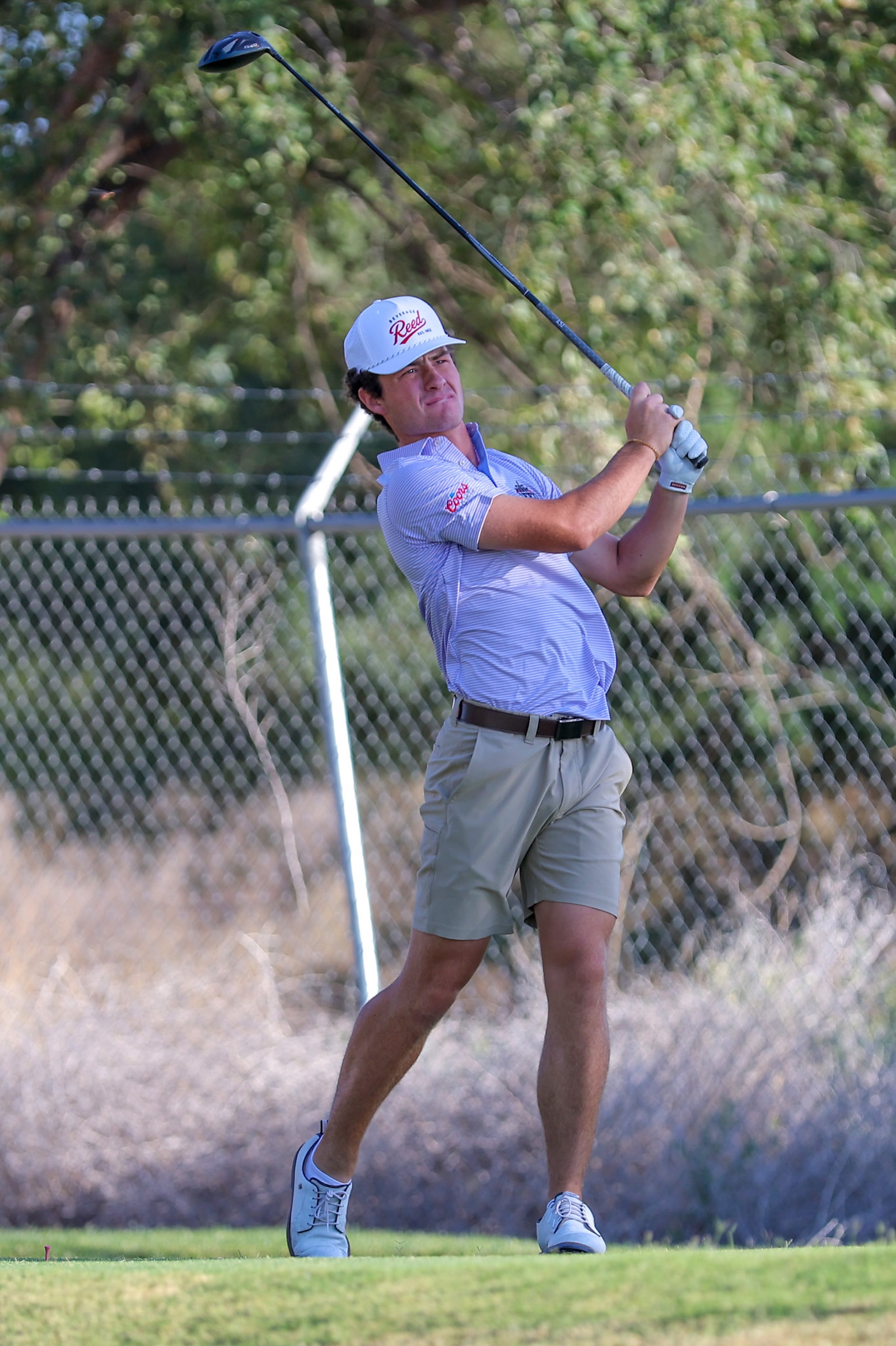 Male golfer in a light striped polo swings a driver on a grassy course, fence and trees in background