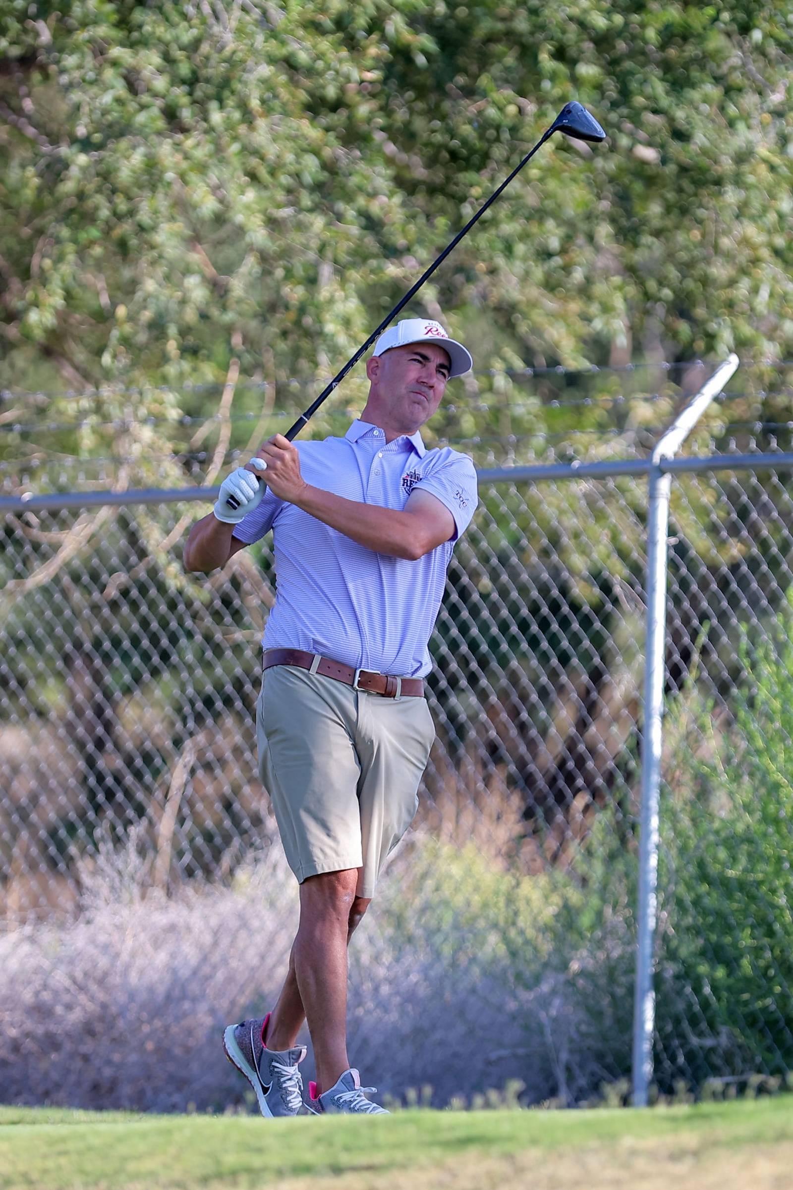 Male golfer in mid-swing on a golf course, wearing a light blue polo, white cap, and khaki shorts.